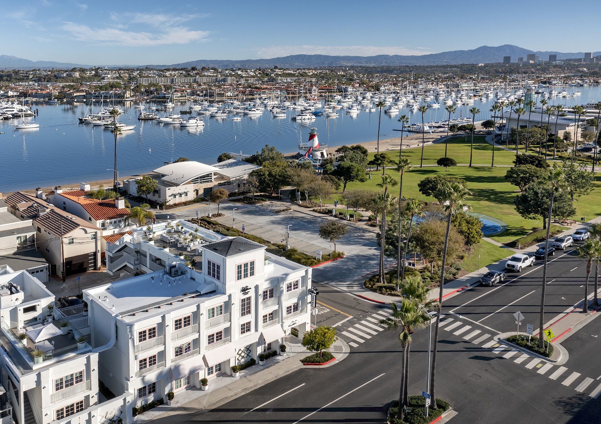 A sunny coastal marina with sailboats, palm trees, a grassy waterfront, and mountains in the background.