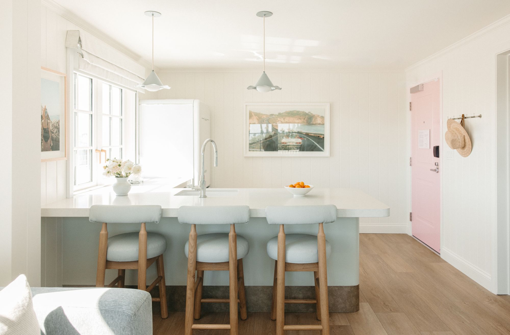 A bright kitchen dining area with a white table, blue stools, pendant lights, a sink, framed wall photo, and a pink door.