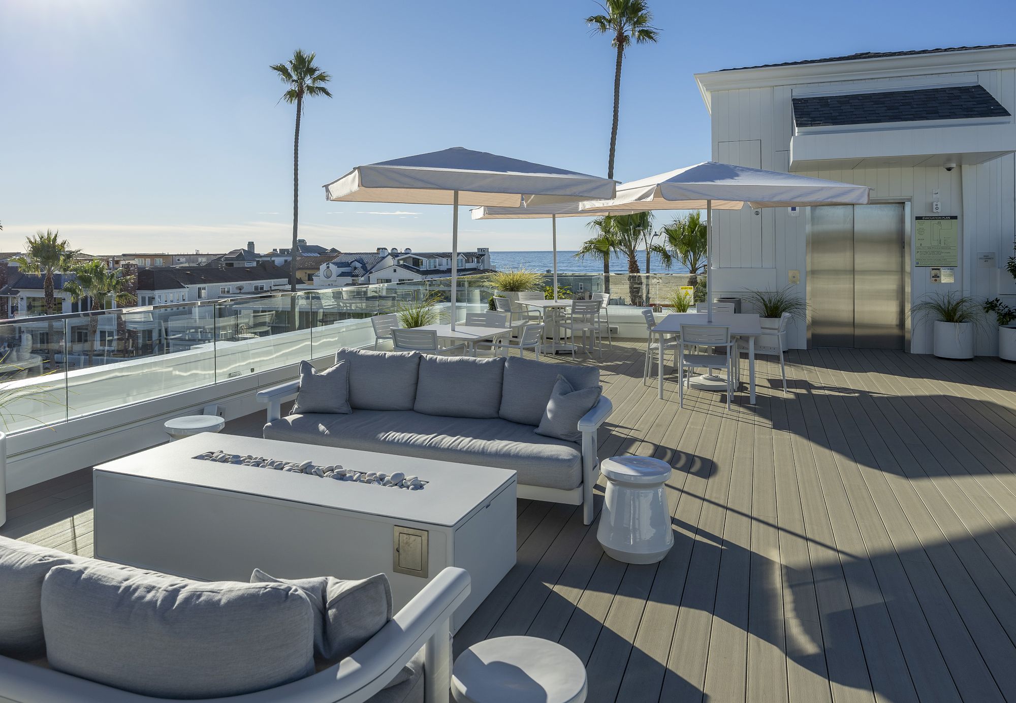 A sunny ocean-view rooftop patio with a gray sofa, dining set, striped umbrellas, palm trees, and a glass railing.