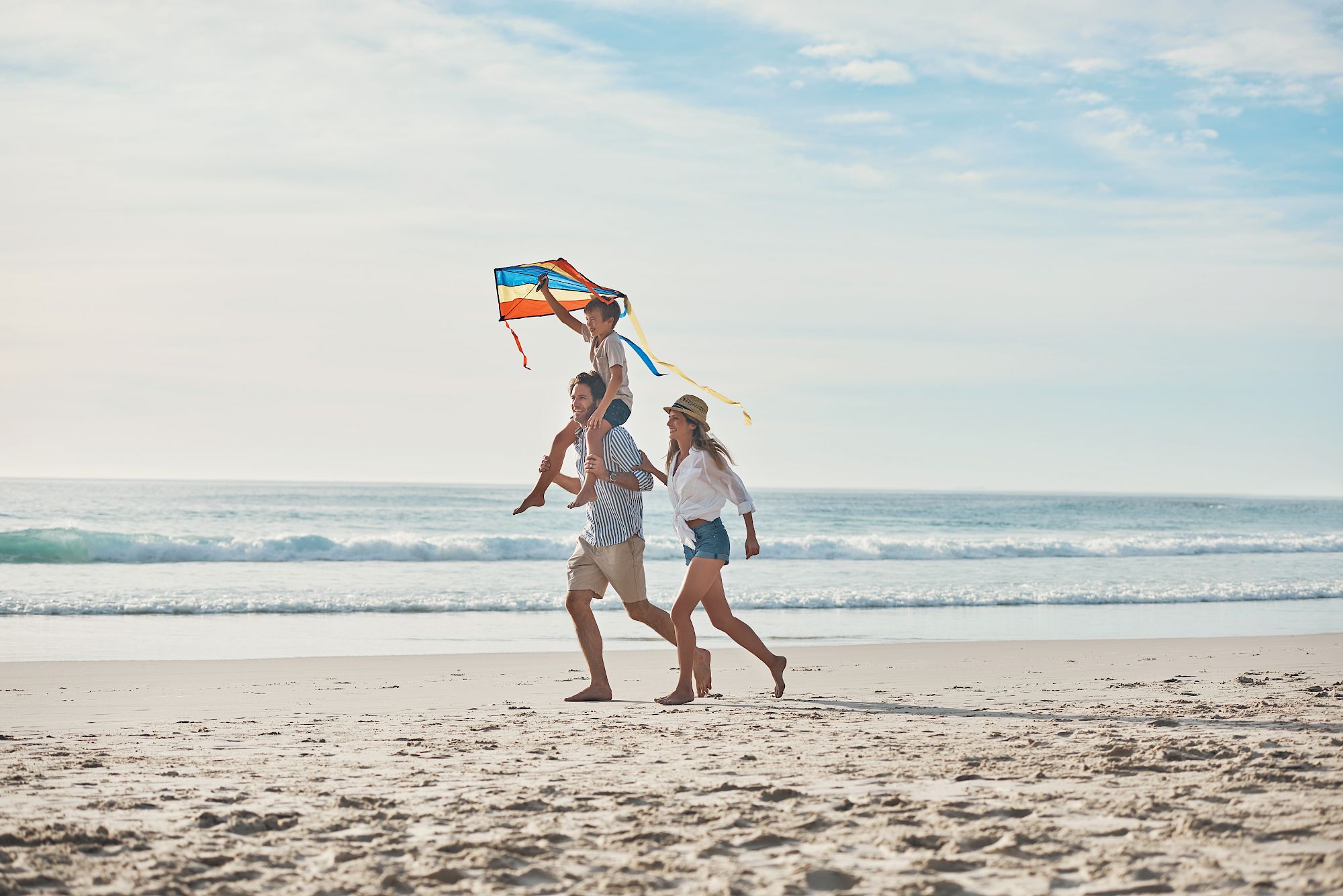A family runs along the beach flying a colorful kite, laughing together under a bright sky.