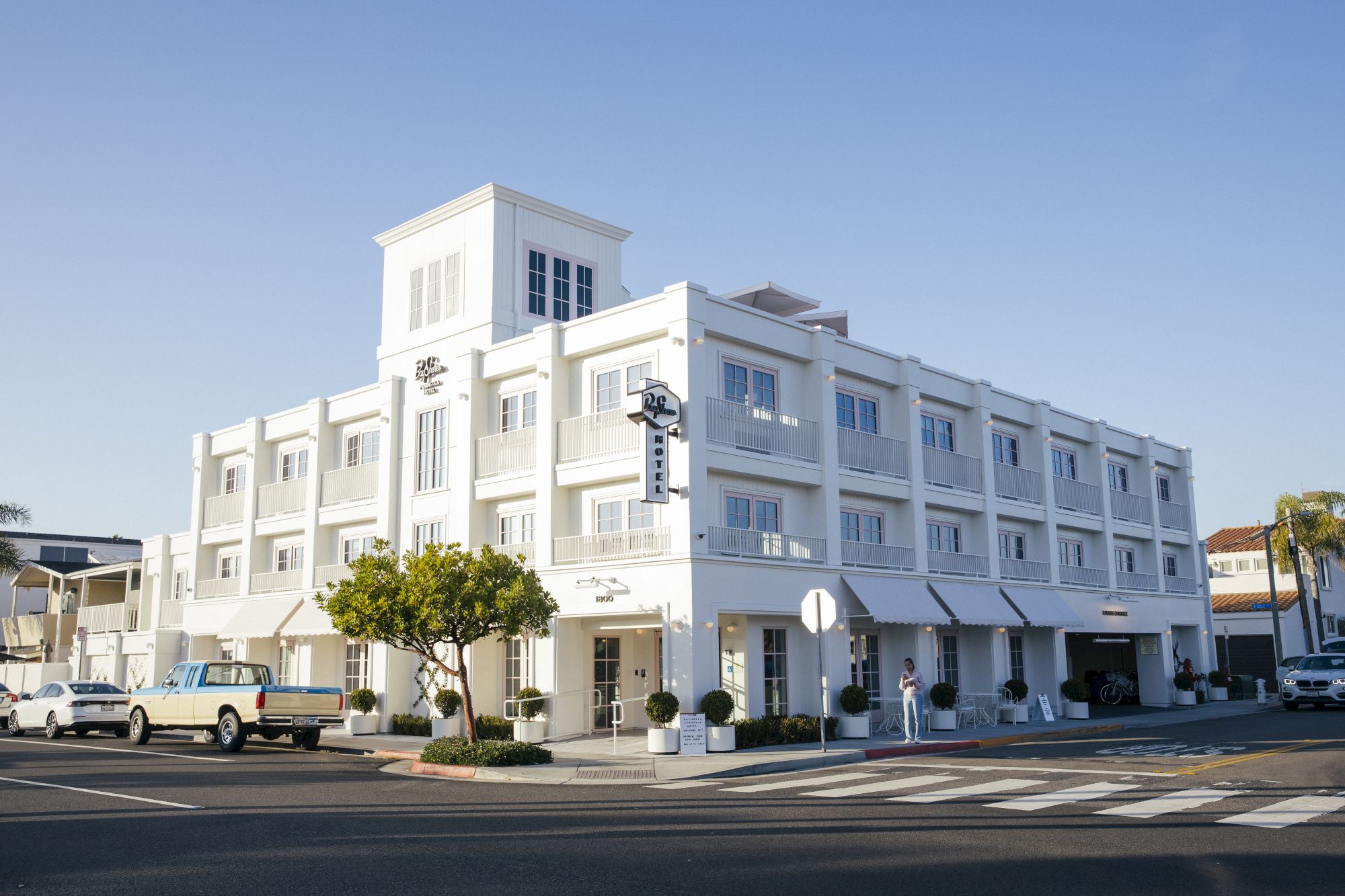 A blue-and-white multi-story hotel building with balconies and parked cars stands under a clear sky.