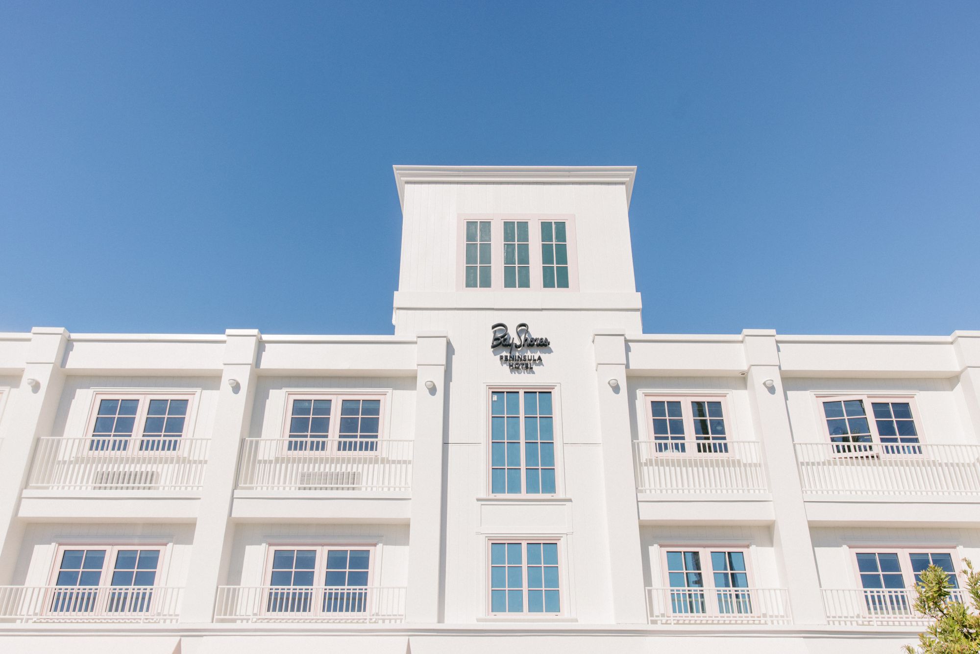 A white hotel building fa&ccedil;ade with the &ldquo;Bay Shores Peninsula&rdquo; sign and blue windows under a clear sky.