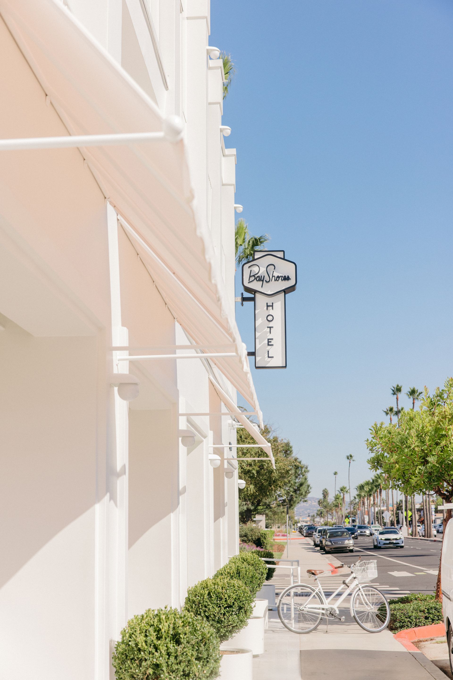 White building facade on a sunny street with a hanging sign reading &ldquo;Bay Shores&rdquo; and a tall &ldquo;HOTEL&rdquo; marker on a pole.