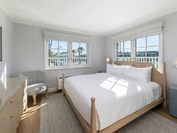 Bright seaside-style bedroom with a wooden bed, white linens, bedside lamps, two large windows, and striped wallpaper.