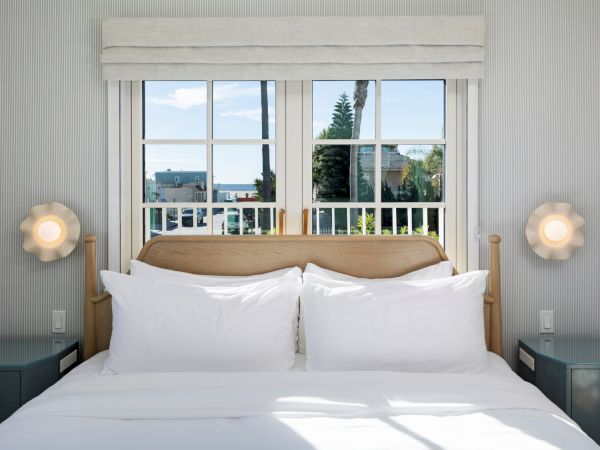 A modern hotel bedroom with a neatly made bed, bedside lights, and a view of palm trees through large windows.