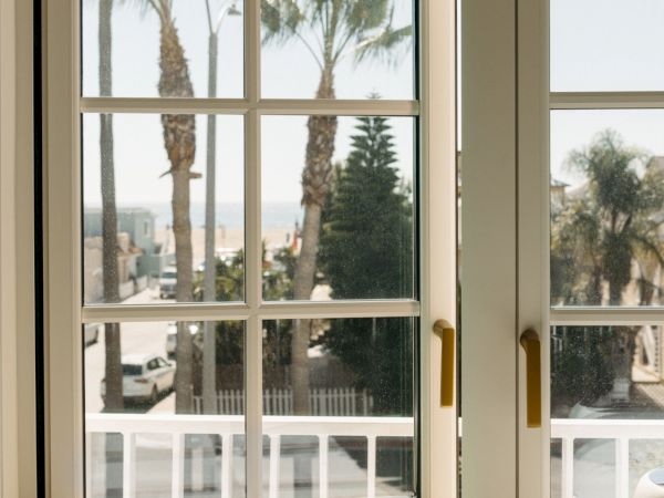 View through a large window with droplets on the glass, palm trees and distant sea beyond, sunlight on the windowsill beside a mug.