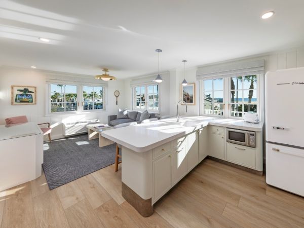 A bright modern kitchen/living room with a white island, pendant lights, large windows, and a glimpse of the ocean outside.