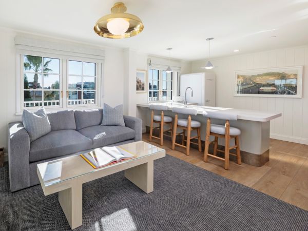 Bright living area with gray sofa and glass coffee table, plus a kitchen island, bar stools, pendant lights, and windows showing a road view.
