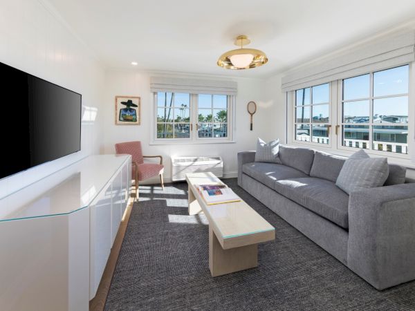 Bright living room with a flat-screen TV, gray sofa, coffee table, and large windows with blinds under a ceiling light.