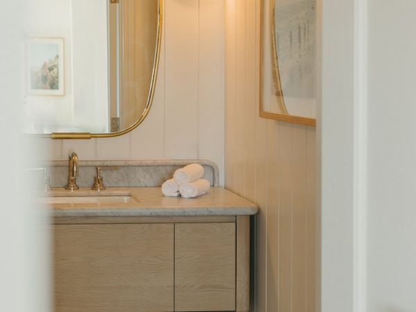 A stylish bathroom vanity with a marble top, stacked white towels, a warm round light, and a curved gold-framed mirror.