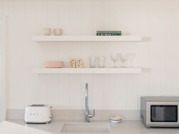 A bright kitchen shelf and counter with mugs, glasses, stacked plates, a kettle/toaster, and a cookbook on display.