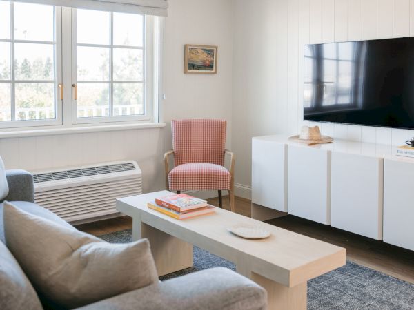Bright living room with a sofa, red-checkered chair, big TV, small books on a cabinet, and large windows showing trees.