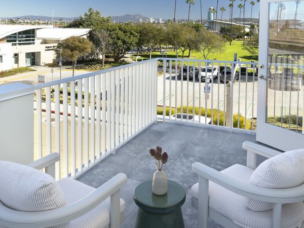 Balcony with cushioned seating overlooking a sunny landscaped campus, palm trees, cars below, and mountains in the distance.