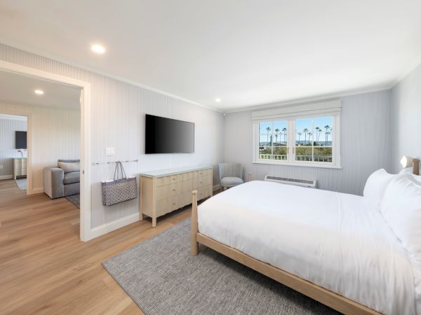 Bright modern bedroom with a bed, mounted TV, dresser, chairs, and large windows showing palm trees outside.