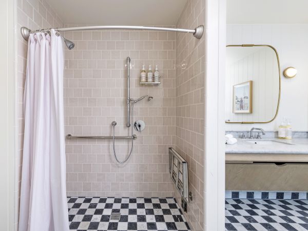 A bright bathroom with a tiled shower, pink curtain, chrome showerhead, and a vanity with mirror, lights, and toiletries.