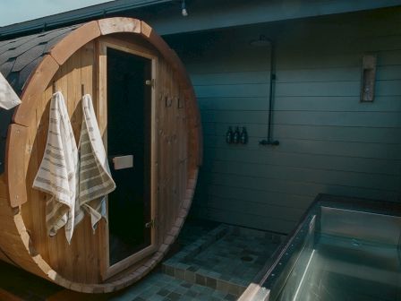 A wooden barrel sauna with a door open, towels hanging inside, blue tiled room, and a bench or stove on the right.