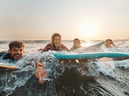 Three friends laugh while riding a teal surfboard through splashing waves at the beach.