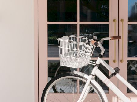 A white bicycle with a front wire basket is parked outside a building by pink-framed windows.