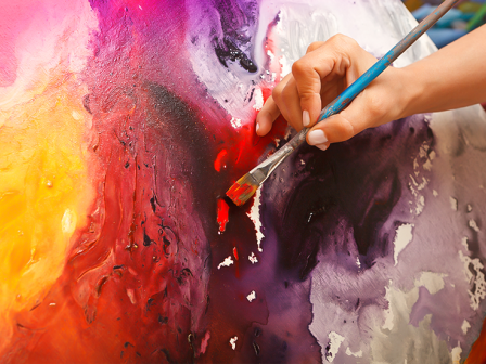 A person paints an abstract, colorful canvas with a brush, blending bright pinks, purples, reds, and yellows into swirling textures.