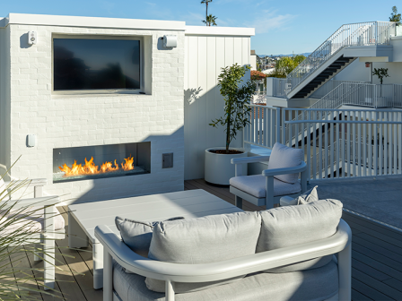 A modern rooftop patio with white lounge furniture, a built-in fireplace, and a blue deck railing under a clear sky.