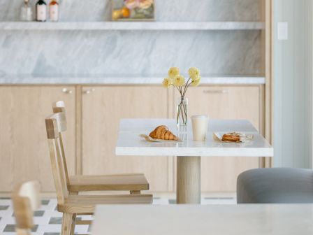 A bright kitchen scene with wooden shelves, bottles and artwork on the wall, flowers by the counter, and pastries on the table.