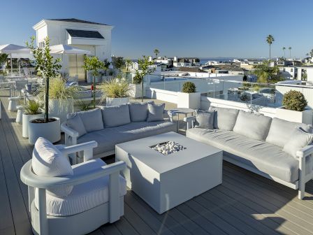 A sunny rooftop patio with a white sectional sofa, coffee table, planters, umbrellas, and a view of homes and distant hills.
