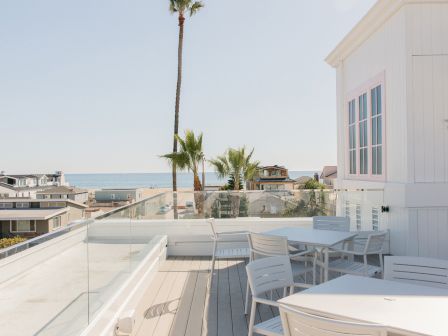 A bright ocean-view rooftop patio with white lounge chairs, a glass railing, and tall palm trees beside a white building.