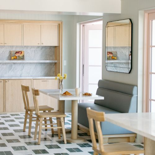 A bright modern kitchen-dining area with wooden cabinets, a small table and chairs, a gray sofa, and a large wall mirror.