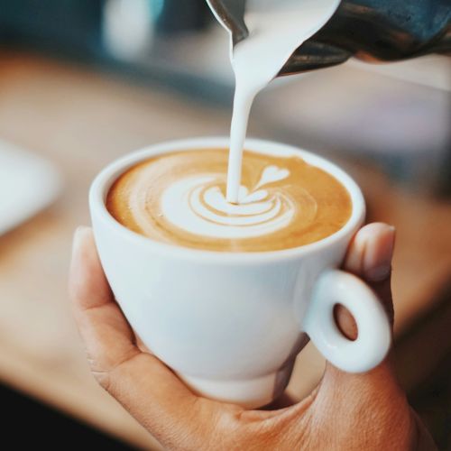 A hand holds a white cup while milk is poured onto coffee, creating latte art in a warm espresso drink.
