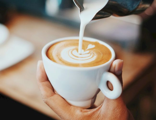 A hand holds a white cup while milk is poured onto coffee, creating latte art in a warm espresso drink.