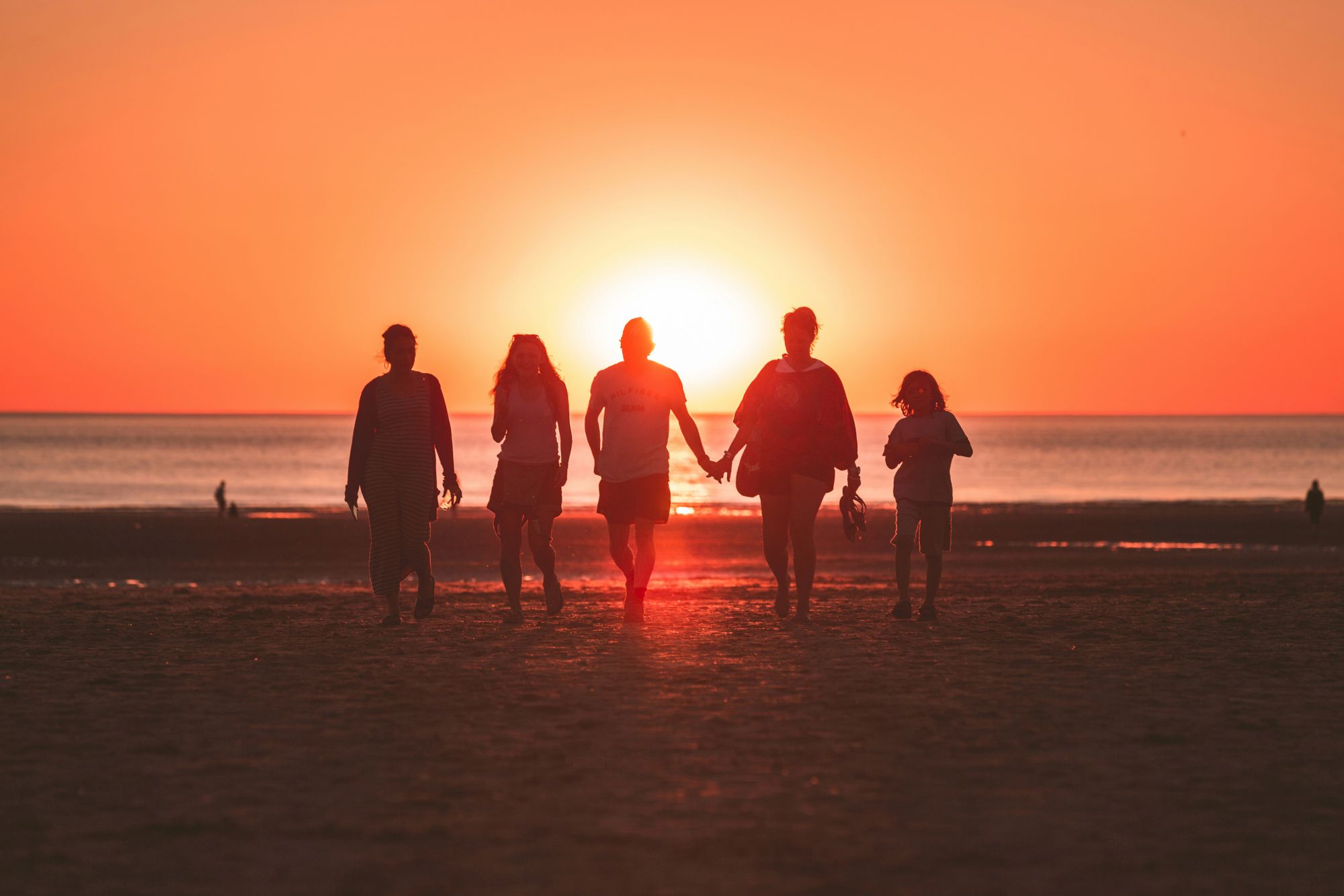 Silhouetted group walking hand-in-hand along a beach at sunset, with the bright sun glowing behind them.
