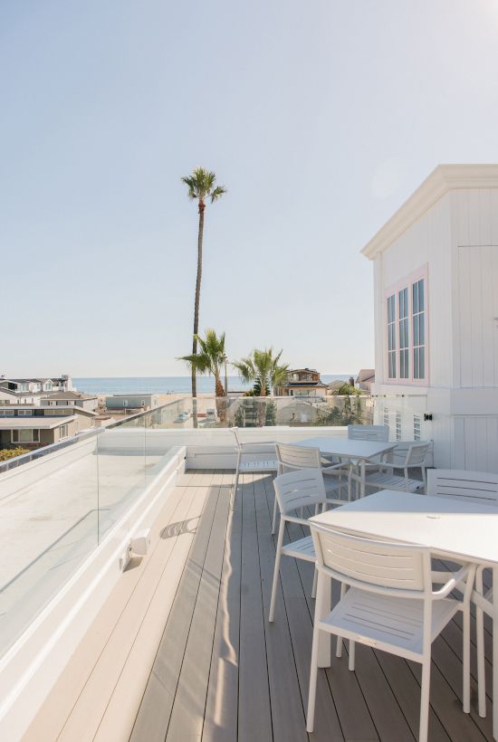 Sunlit oceanfront rooftop terrace with white chairs, glass railing, palm tree, and a modern beachfront house under a clear sky.