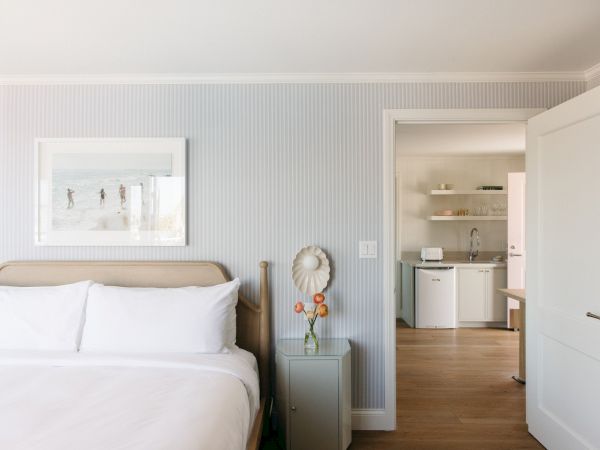 A bright bedroom with a bed, framed beach print, and a doorway to a small kitchenette with shelves, sink, and appliances.