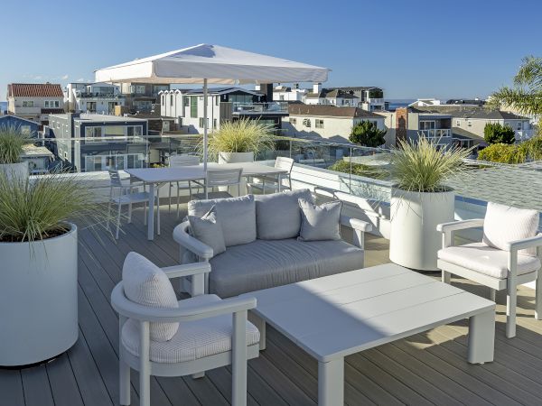 A sunny rooftop deck with outdoor seating and planters overlooking a coastal neighborhood and distant sea views.