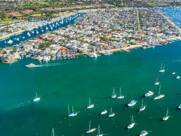 Aerial view of a coastal town and marina packed with sailboats, with city buildings and mountains in the background.