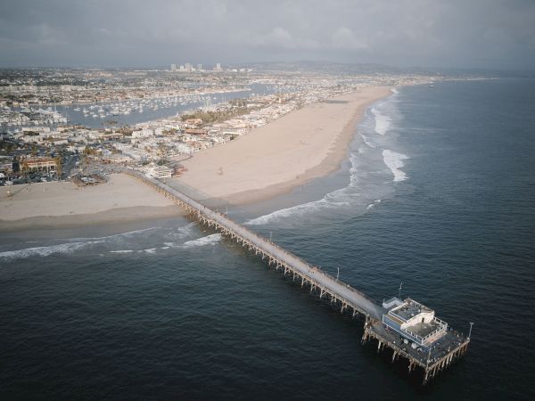 Aerial view of a wide sandy beach with a long pier and bustling harbor, overlooking a coastal town and marina boats.
