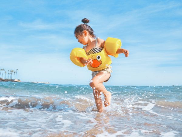 A little girl splashes in the ocean, wearing a yellow cartoon float ring on a sunny beach day.