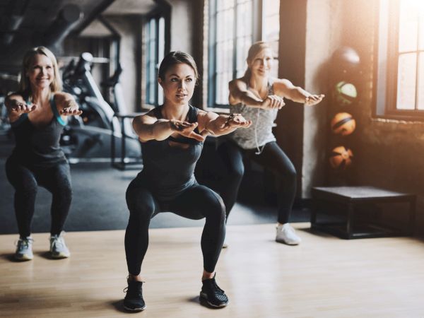Three women in a gym lead a group workout, lunging with arms extended while others mirror behind them.