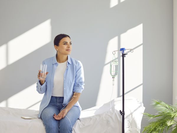 A woman on a bed holds a glass of water while an IV drip runs beside her in a bright room.