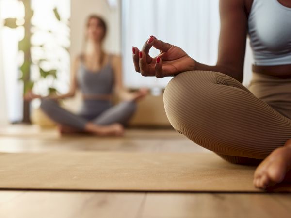 A person&rsquo;s hands form a yoga gesture in the foreground while another person meditates on a mat in soft, bright light.
