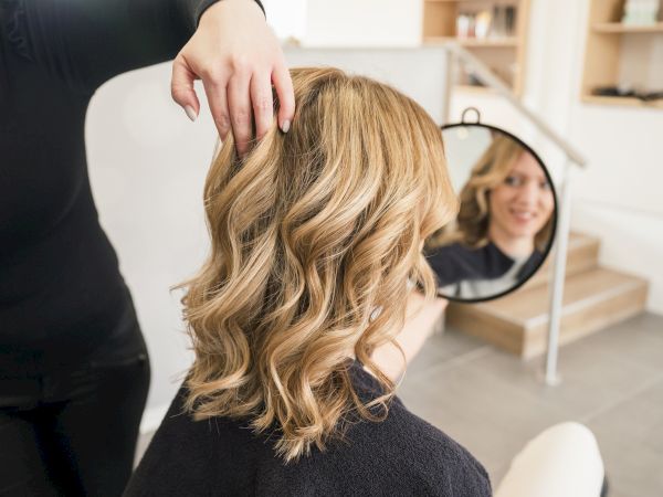 A stylist gently styles a woman&rsquo;s long blonde curls in a salon while she watches her reflection in a mirror.