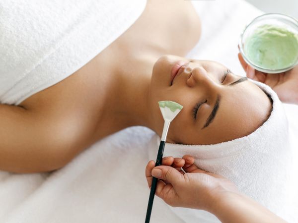 A spa facial: a person lies back while a beautician applies a green mask with a brush, holding a jar nearby.