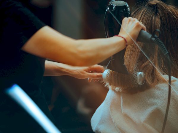 A hairstylist blow-dries and styles a woman&rsquo;s long hair with a round brush and hair dryer.