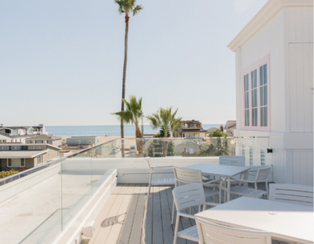 Seaside balcony with glass railing, white chairs/tables, palm trees, and an ocean view beside coastal houses.