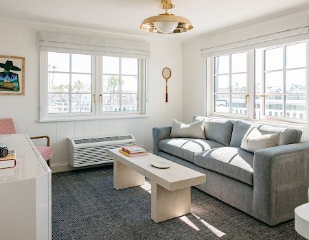 A bright living room with a grey sofa, coffee table, books, wall art, and large windows with white shades.
