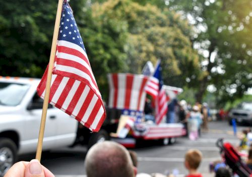 A hand holds a waving American flag while a crowd watches a parade with more U.S. flags along the street.