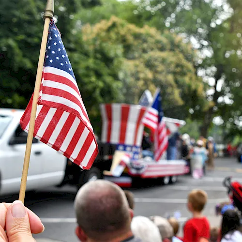 A hand holds a waving American flag while a crowd watches a parade with more U.S. flags along the street.