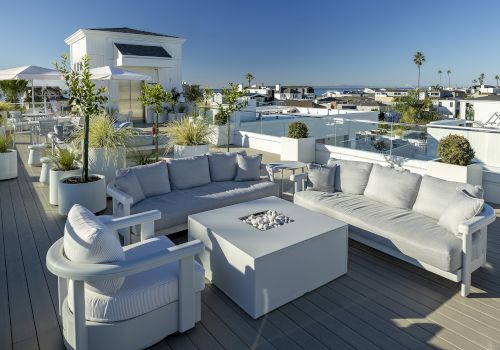 A bright rooftop patio with white outdoor sofas, a firepit table, potted plants, and a city view under a clear blue sky.
