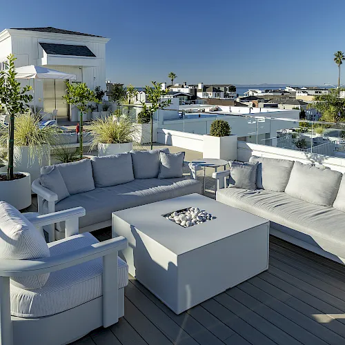 A bright rooftop patio with white outdoor sofas, a firepit table, potted plants, and a city view under a clear blue sky.