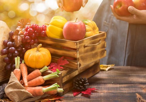 A hand reaches toward apples while an autumn harvest of fruits and vegetables sits in wooden crates on a rustic table.